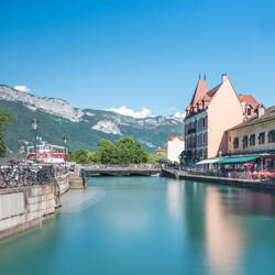 Annecy : Canal paisible traversant Annecy avec des bâtiments historiques aux façades claires et aux toits rouges bordant ses rives, un pont au centre, des montagnes imposantes en arrière-plan sous un ciel bleu clair.
