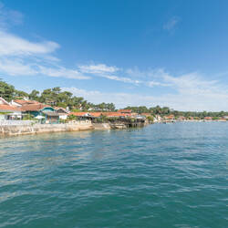 Arcachon : Village côtier avec des maisons aux toits rouges bordant une étendue d'eau bleue ondulante sous un ciel bleu avec quelques nuages blancs.