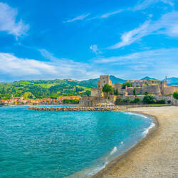 Argeles sur Mer : Vue d'une plage de galets bordant une mer turquoise, avec un château médiéval en pierre dominant le rivage, une ville colorée en arrière-plan et des collines verdoyantes sous un ciel bleu avec des nuages.