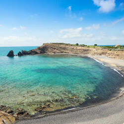 Cap d'Agde : Crique isolée avec une plage de sable sombre bordée d'une eau turquoise claire révélant des rochers, entourée de falaises rocheuses et d'une végétation clairsemée sous un ciel bleu avec quelques nuages.