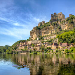Dordogne : Village médiéval au bord d'une rivière calme, dominé par un château perché sur une falaise abrupte sous un ciel bleu partiellement nuageux.