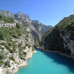 Gorges du Verdon avec une rivière turquoise serpentant entre d'imposantes falaises rocheuses et une végétation verdoyante sous un ciel bleu clair.
