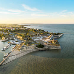 Ile d'Oléron : Vue aérienne de la citadelle de Saint-Martin-de-Ré, une forteresse en étoile avec des remparts entourant une partie de la ville côtière et son port rempli de bateaux, bordée par la mer sous un ciel clair au crépuscule.