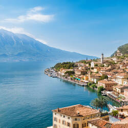 Italie : Vue panoramique de Limone sul Garda, une ville pittoresque avec des maisons aux toits rouges le long du lac de Garde bleu, avec des montagnes imposantes en arrière-plan sous un ciel clair.