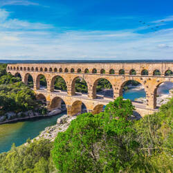 Languedoc-Roussillon : Vue du Pont du Gard, un ancien aqueduc romain à trois niveaux avec de multiples arches en pierre ocre, traversant une rivière turquoise bordée d'une végétation luxuriante sous un ciel bleu avec quelques nuages.