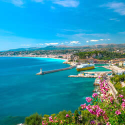 Côte d'Azur : Vue panoramique du port de Nice avec un grand ferry jaune, un phare sur une jetée, des montagnes en arrière-plan et des fleurs roses au premier plan, sous un ciel bleu clair.