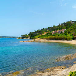 Var : Crique isolée avec une plage de sable clair bordée d'une eau turquoise et de rochers, avec une colline verdoyante parsemée de maisons sous un ciel bleu serein.