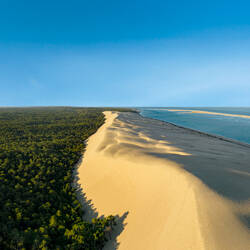 Les Landes : Vue aérienne de la Dune du Pilat, une immense dune de sable beige bordée d'une forêt de pins verts d'un côté et de l'océan bleu clair de l'autre, sous un ciel bleu uniforme.