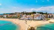 Vue aérienne de la Platja de Blanes en Espagne, montrant deux plages de sable doré séparées par une formation rocheuse et une jetée, bordées par des bâtiments colorés sous un ciel bleu parsemé de quelques nuages.