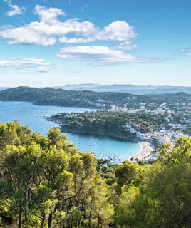 Vue aérienne de la côte de Begur sur la Costa Brava, avec une plage de sable en demi-lune, de nombreux bateaux dans la baie, et des maisons blanches entourées de végétation verdoyante, sous un ciel bleu parsemé de nuages blancs.