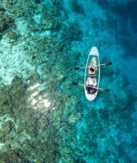Vue aérienne d'un kayak transparent avec deux personnes pagayant sur des eaux turquoise claires, révélant les fonds marins rocailleux. Le lieu n'est pas identifiable mais pourrait être une crique de la Costa Dorada.