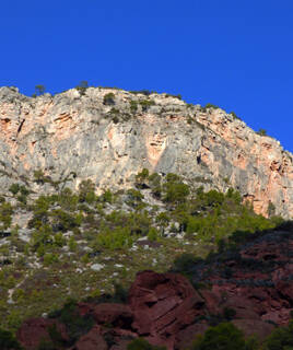 Sommet du Parc Naturel de Montsant, avec des falaises rocheuses aux teintes grises et ocre, et des pins dispersés sur ses pentes, sous un ciel bleu intense. Au premier plan, des roches rouges.