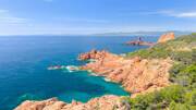 Paysage côtier ensoleillé du Cap Dramont à Saint-Raphaël, montrant les roches rouges du Massif de l'Esterel plongeant dans la mer Méditerranée turquoise, avec une tour visible au loin sur un îlot rocheux.