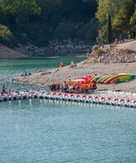 Des baigneurs profitent des eaux turquoise du Lac de Sainte-Croix, au cœur des Gorges du Verdon, avec des falaises et une végétation luxuriante en arrière-plan, sous un grand ciel bleu.