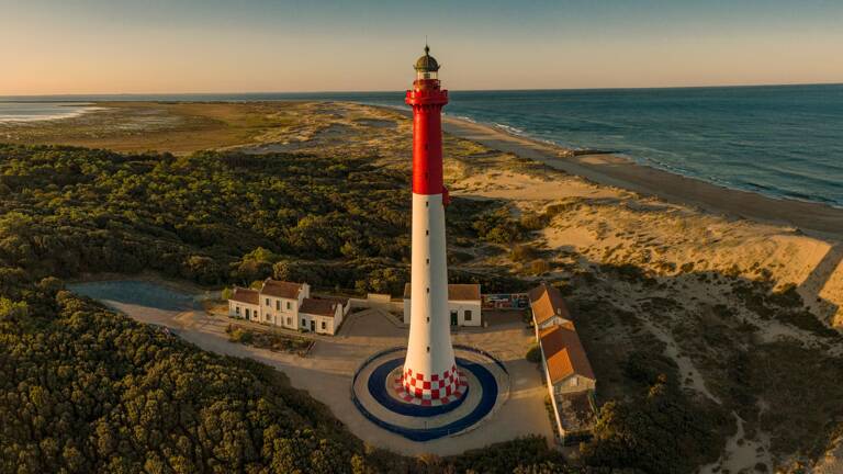 Vue aérienne du Phare des Baleines à Saint-Clément-des-Baleines, sur l'Île de Ré, avec sa tour rouge et blanche caractéristique, entouré de dunes de sable, de végétation dense et de l'océan Atlantique au coucher du soleil.