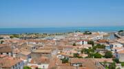 Vue aérienne du village de Saint-Martin-de-Ré sur l'Île de Ré, montrant un ensemble de toits de tuiles rouges, le port avec ses bateaux et l'océan Atlantique en arrière-plan sous un ciel bleu clair.
