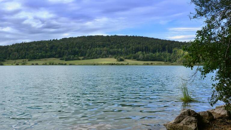Vue sur le paisible lac du Vouglans avec une forêt et une colline en arrière-plan sous un ciel nuageux.