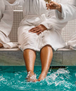 Pieds éclaboussant l'eau d'une piscine aux Thermes de Lons-le-Saunier, avec des personnes en peignoir tenant des verres de champagne.