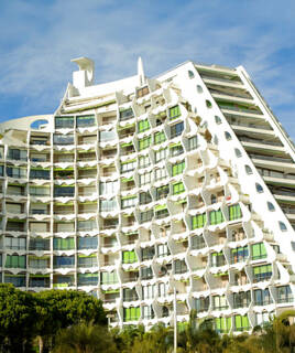 Bâtiment à l'architecture pyramidale emblématique de La Grande Motte, avec des balcons ondulants et des touches de vert, sous un ciel bleu.