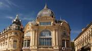 Un majestueux bâtiment haussmannien avec une grande coupole et des ornements, sous un ciel bleu éclatant, situé sur la Place de la Comédie à Montpellier.