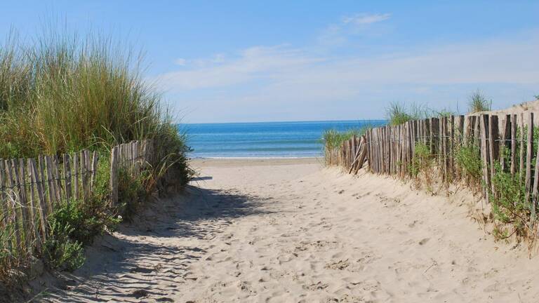 Allée sablonneuse bordée de clôtures en bois et de végétation menant à la Plage du Grand Travers à La Grande Motte, sous un ciel bleu.