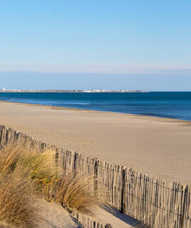 Plage de sable doré de La Grande Motte, bordée par une clôture en bois et de l'herbe sèche, avec la mer calme et l'horizon dégagé sous un ciel clair.