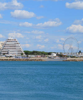 Une vue de La Grande-Motte avec la mer au premier plan, un bâtiment pyramidal emblématique et une grande roue en arrière-plan sous un ciel bleu parsemé de nuages blancs.
