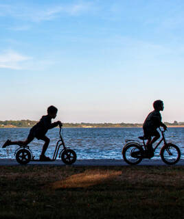 Silhouette de deux enfants, l'un sur une trottinette et l'autre sur un vélo, longeant la côte sur la piste cyclable de la Vélodyssée à La Tranche-sur-Mer, avec la mer en arrière-plan au coucher du soleil.
