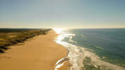 Vue aérienne d'une longue plage de sable doré et de l'océan Atlantique à Lacanau, avec des dunes recouvertes d'herbe sur la gauche et le soleil scintillant sur l'eau à l'horizon.