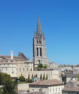 La haute flèche de l'Église Monolithe de Saint-Émilion domine les toits des bâtiments historiques de la ville, sous un ciel d'un bleu éclatant.