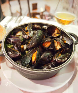Une généreuse portion de mouclade vendéenne, plat typique de Longeville-sur-Mer, servie dans une marmite noire, avec des moules cuites à la vapeur dans une sauce, accompagnée d'un verre de boisson en arrière-plan.