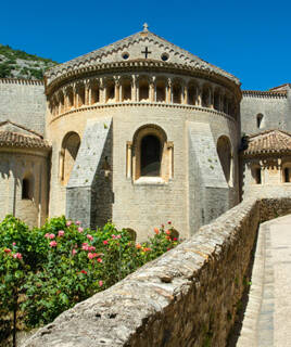 Vue de l'arrière de l'Abbaye de Saint-Guilhem-le-Désert, un village médiéval près de Marseillan-Plage, montrant son architecture romane en pierre, des murs courbes et des arcades, avec des roses en fleurs au premier plan et des montagnes en arrière-plan.