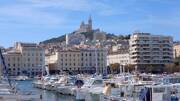 Vue emblématique du Vieux-Port de Marseille, avec ses nombreux bateaux amarrés au premier plan et la ville en arrière-plan, dominée par la basilique Notre-Dame de la Garde perchée sur une colline sous un ciel bleu.