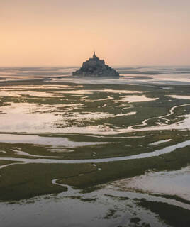 Le Mont Saint-Michel au loin, entouré par l'étendue de sable et les méandres verdoyants des marais, sous un ciel pâle au lever ou au coucher du soleil.