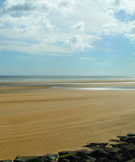 Vaste étendue de sable doré sur Juno Beach en Normandie, avec la mer calme et un ciel parsemé de nuages, et quelques rochers au premier plan.
