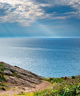 Vue panoramique de la mer à la Pointe du Grouin en Bretagne, avec des falaises rocheuses couvertes de végétation au premier plan et un ciel nuageux laissant passer des rayons de soleil.