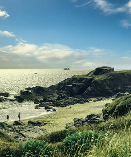 Une crique sauvage sur la côte du Morbihan, avec des rochers, une petite plage et des personnes au bord de l'eau, sous un ciel partiellement nuageux. Au loin, on aperçoit un bateau sur la mer.