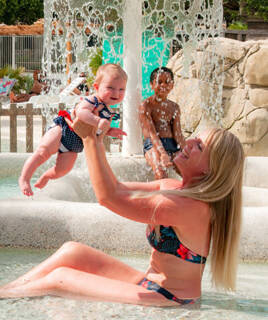 Femme souriante tenant un bébé au-dessus de l'eau dans une pataugeoire, avec un enfant en arrière-plan.