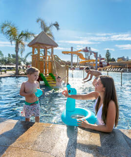 Famille s'amusant dans une pataugeoire d'un parc aquatique avec toboggans et jeux, probablement dans un camping ou un complexe de loisirs.