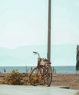 Un vélo rouge vintage garé près de la plage, avec la mer et des montagnes en arrière-plan sous un ciel clair, évoquant l'esprit de la Vélodyssée.