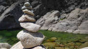 Un empilement de pierres (cairn) se dresse en équilibre au bord d'une piscine naturelle aux eaux vertes et translucides, creusée dans la roche près de Porto-Vecchio.