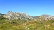Paysage de montagne des Pyrénées-Atlantiques avec des sommets rocheux, des pâturages verts parsemés de troupeaux, et un chemin menant à quelques bâtiments isolés, situé dans le Parc National des Pyrénées.