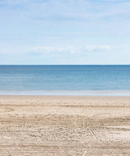 La vaste plage de sable de L'Aiguillon-sur-Mer, avec les traces de véhicules sur le sable, s'étendant jusqu'à l'océan calme sous un ciel bleu clair parsemé de nuages blancs.