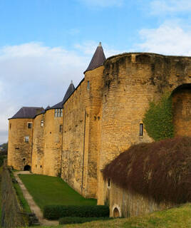 Façade extérieure en pierre du Château de Sedan dans les Ardennes, avec des tours pointues et un mur d'enceinte couvert de végétation, sous un ciel nuageux.