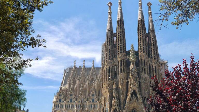 La basilique de la Sagrada Familia à Barcelone, avec ses tours majestueuses et sa façade détaillée, vue à travers la verdure luxuriante de l'environnement, sous un ciel bleu parsemé de nuages blancs.