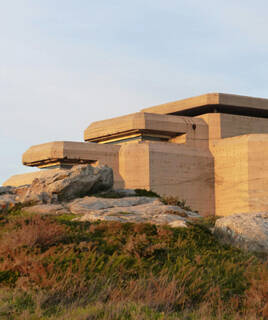 Le Grand Blockhaus de Batz-sur-Mer, une structure massive en béton, s'élève sur un terrain rocheux et herbeux sous un ciel clair, éclairé par la lumière du soleil couchant.