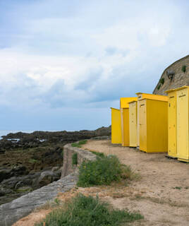 Une rangée de cabines de plage jaunes sur la côte rocheuse de Batz-sur-Mer, sous un ciel nuageux, avec l'océan et ses rochers visibles à gauche.