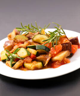 Une assiette de ratatouille, spécialité de la cuisine méditerranéenne de Cagnes-sur-Mer, garnie de légumes du soleil coupés en dés (courgettes, tomates, aubergines) et de brins de romarin frais.