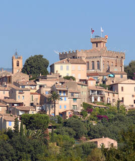Le village médiéval du Haut de Cagnes-sur-Mer, avec le Château Grimaldi dominant les maisons aux toits de tuiles et aux volets colorés, entouré de végétation sous un ciel bleu clair.