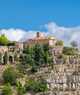 Le village perché de Gourdon, près de Cannes, avec ses bâtiments en pierre et son église à clocher, s'accrochant à une falaise rocheuse escarpée, entouré de verdure sous un ciel bleu parsemé de nuages blancs.